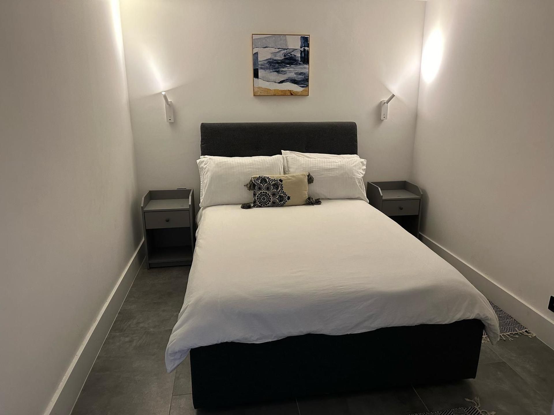 Un dormitorio con una cama, dos mesitas de noche, una lámpara de pared y un cuadro sobre la cama.
