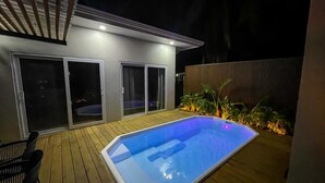 Une piscine avec une terrasse en bois, un patio couvert et une porte-fenêtre coulissante donnant sur un espace extérieur abrité.