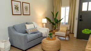 Un salon avec un canapé gris, un pouf rond en fibres tressées, une petite table sur laquelle est posée une plante, et un fauteuil avec un coussin.