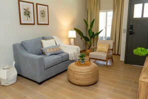 Una sala de estar con un sofá gris, un otoman tejido redondo, una mesita con una planta y un sillón con cojín.