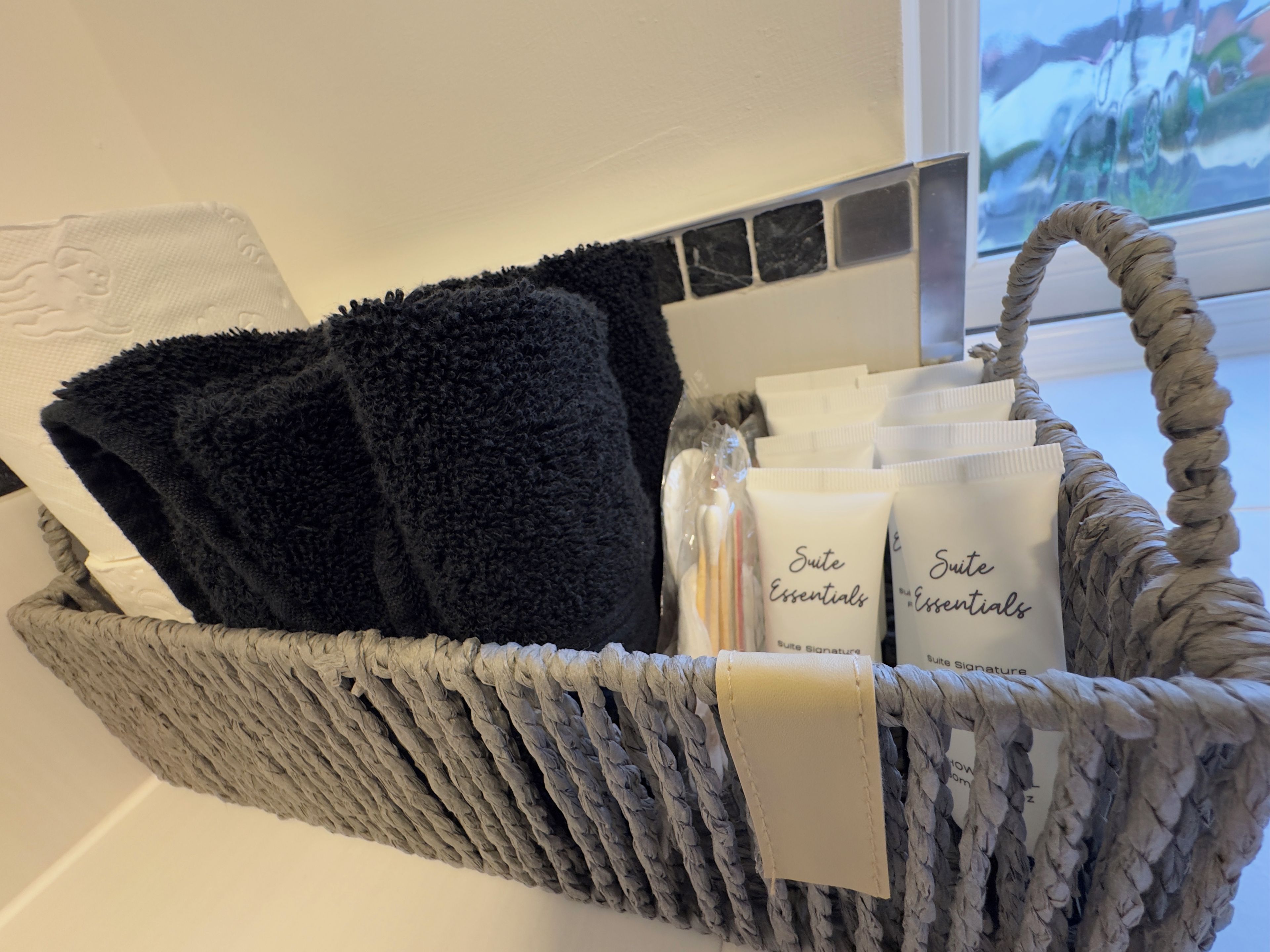 Un panier en osier contenant des serviettes noires, des articles de toilette et un tapis de bain blanc.