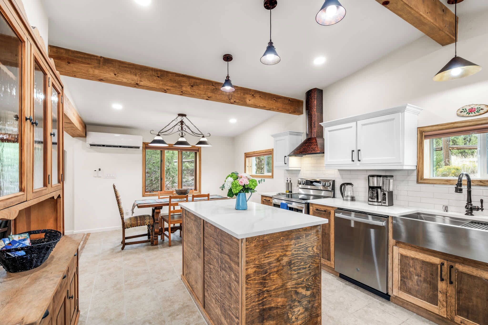 A modern kitchen with wooden beams, a central island, stainless steel appliances, and a dining area with a view of the outdoors.