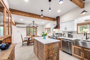 A modern kitchen with wooden beams, a central island, stainless steel appliances, and a dining area with a view of the outdoors. - Renovated Horse Corral Hideaway (Santa Fe)