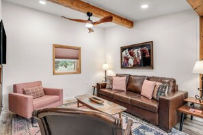 Ein Wohnzimmer mit einem braunen Ledersofa, einem rosafarbenen Sessel, einem Holztisch und einem Deckenventilator.