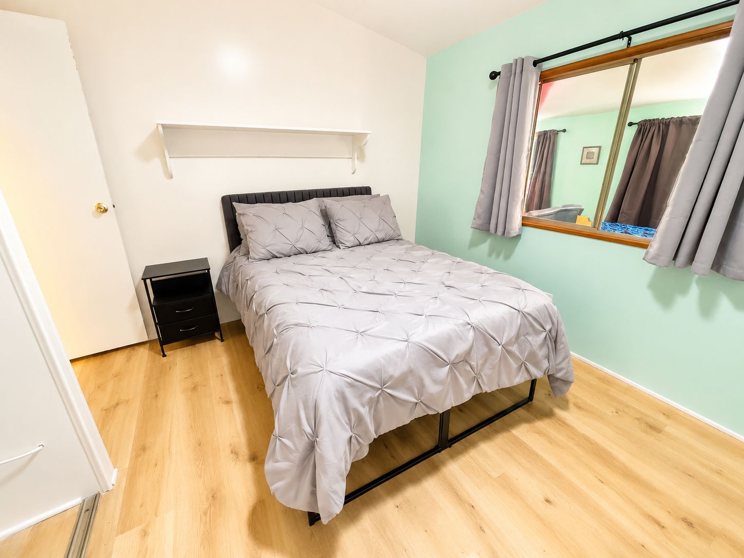 A bedroom with a bed, a nightstand, a shelf, and a window with curtains.