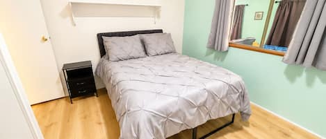 A bedroom with a bed, a nightstand, a shelf, and a window with curtains.