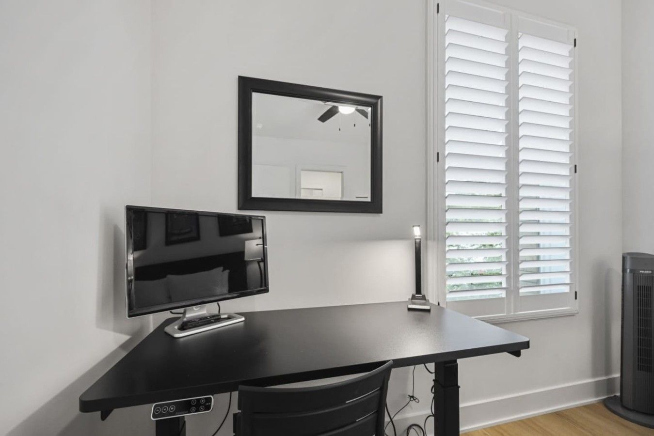 A room with a black desk, a flat-screen TV, a ceiling fan, and a window with blinds.