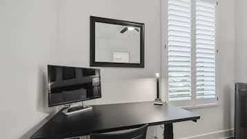 A room with a black desk, a flat-screen TV, a ceiling fan, and a window with blinds.
