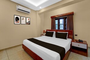 A hotel room with a bed, bedside tables, an air conditioner, a window with curtains, and wall art.