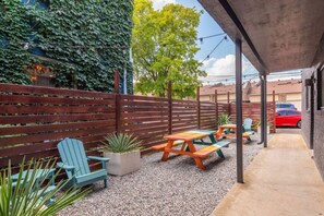 Eine Außenpatio mit Holztischen und -stühlen, einem Betonweg und einem roten Auto im Hintergrund.