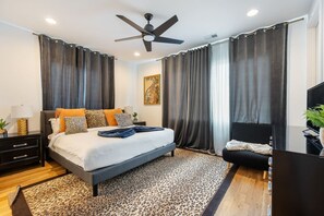 A modern bedroom with a bed, a ceiling fan, a TV, and a large window with curtains. - Last Call Lounge | Rooftop Skyline + Game Room (Nashville)