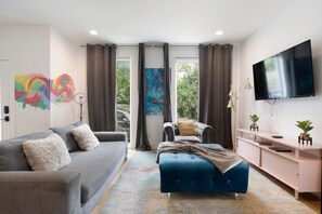 A modern living room with a grey sofa, a blue ottoman, a pink TV stand, and a large TV. - Last Call Lounge | Rooftop Skyline + Game Room (Nashville)