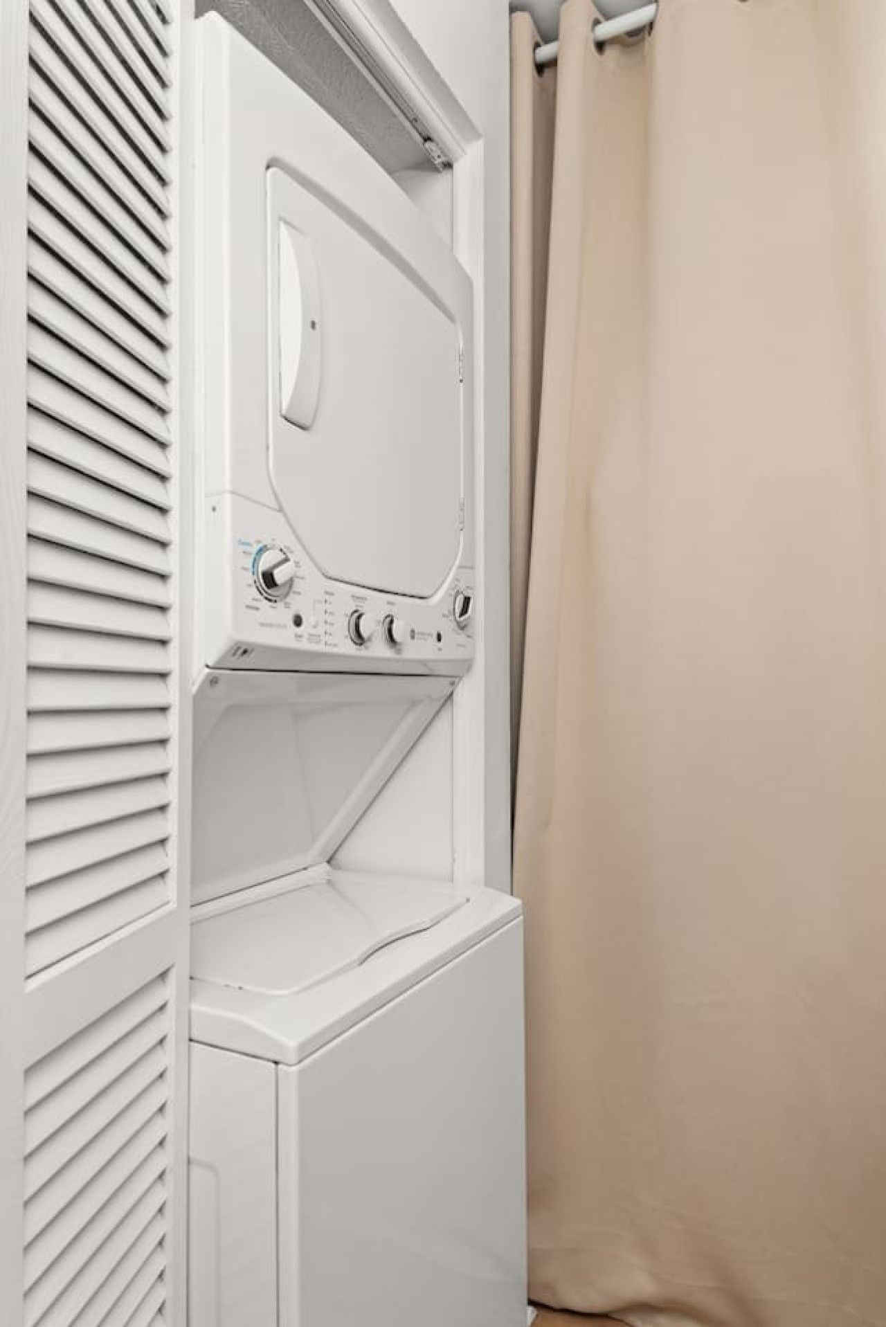 A white laundry room with a dryer, a control panel, and a beige curtain.