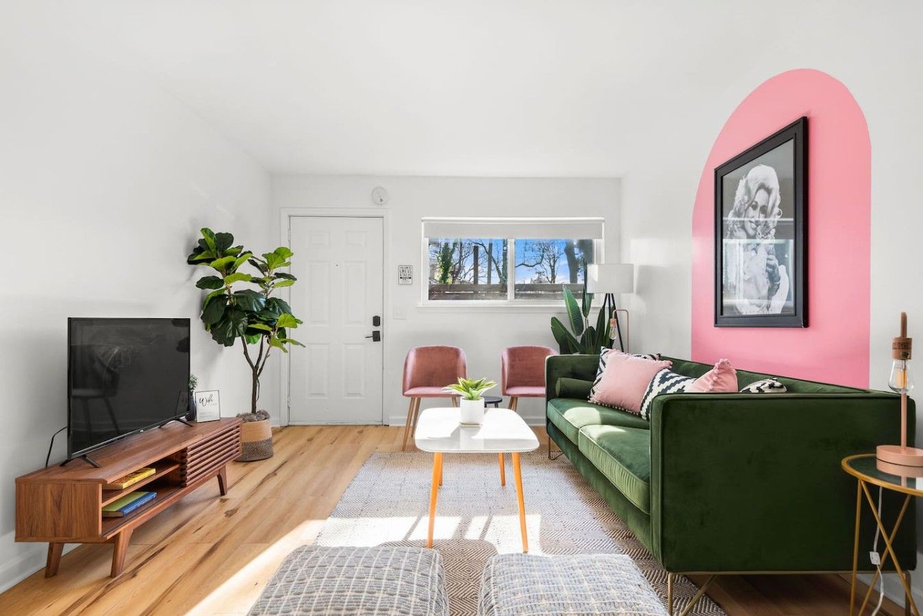 Ein modernes Wohnzimmer mit Fernseher, einem grünen Sofa, einer rosafarbenen Wand und einem großen Fenster.