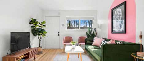 Ein modernes Wohnzimmer mit Fernseher, einem grünen Sofa, einer rosafarbenen Wand und einem großen Fenster.