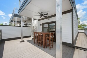 Ein überdachter Außenbereich mit Tisch und Stühlen, einem Deckenventilator und Blick auf ein Gebäude mit einer Wendeltreppe.