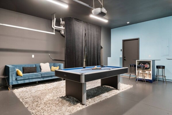 Een pooltafel met een blauwe viltkleed, een blauwe bank met kussens, en een barruimte met barkrukken en een televisie.