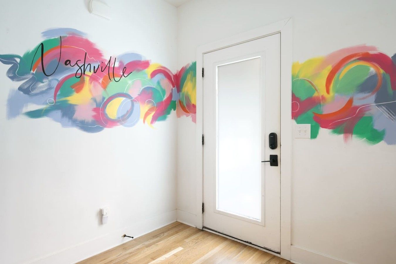 Une pièce avec une porte blanche, un mur orné d’œuvres d’art abstraites aux couleurs vives, et un plancher en bois.