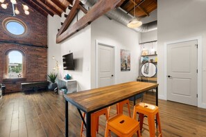 A dining area with a wooden table and orange stools, a brick wall, a TV mounted on the wall, and a window with a view. - Suite and Speakeasy | Pool + Mins to Broadway (Nashville)