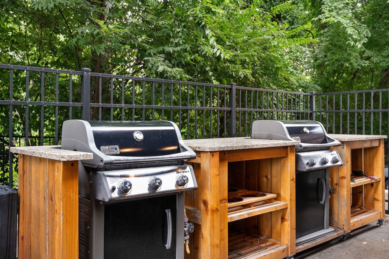 Two outdoor barbecue grills with a wooden structure, a metal fence, and dense foliage in the background.
