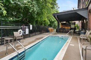 A rectangular pool with a metal ladder, surrounded by a fence, with outdoor seating and a covered bar area in the background. - Suite and Speakeasy | Pool + Mins to Broadway (Nashville)