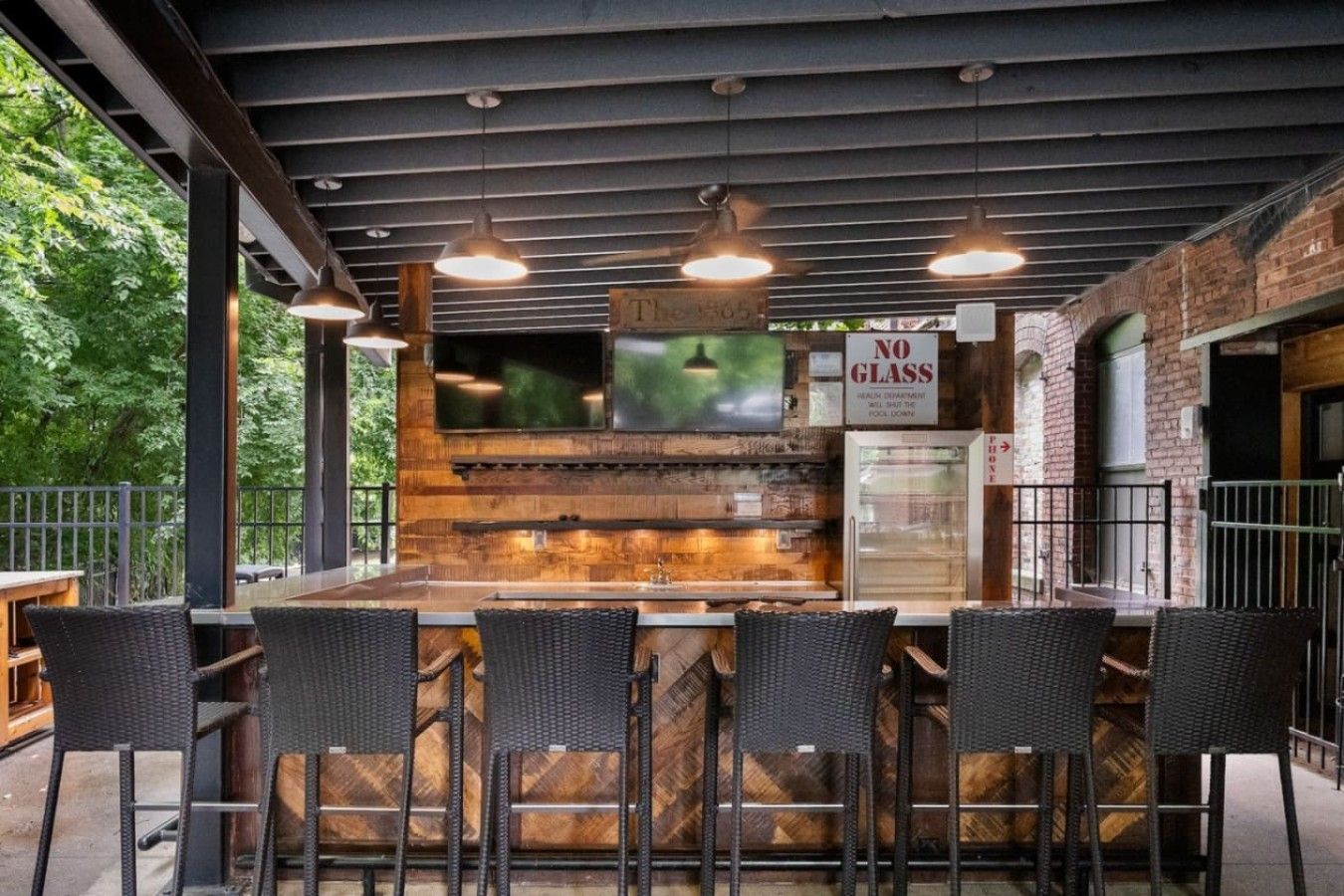 Un espace bar extérieur doté d’un comptoir en bois, de quatre chaises en osier et de luminaires suspendus.