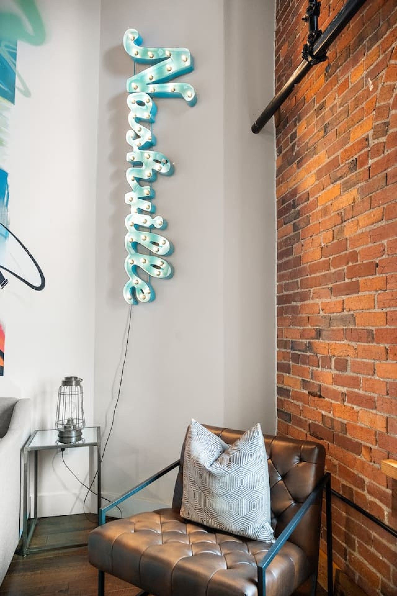 Une pièce avec un fauteuil en cuir, un panneau lumineux au néon qui épelle le mot « Nashville », un mur en briques et une lampe en métal posée sur une table d’appoint.