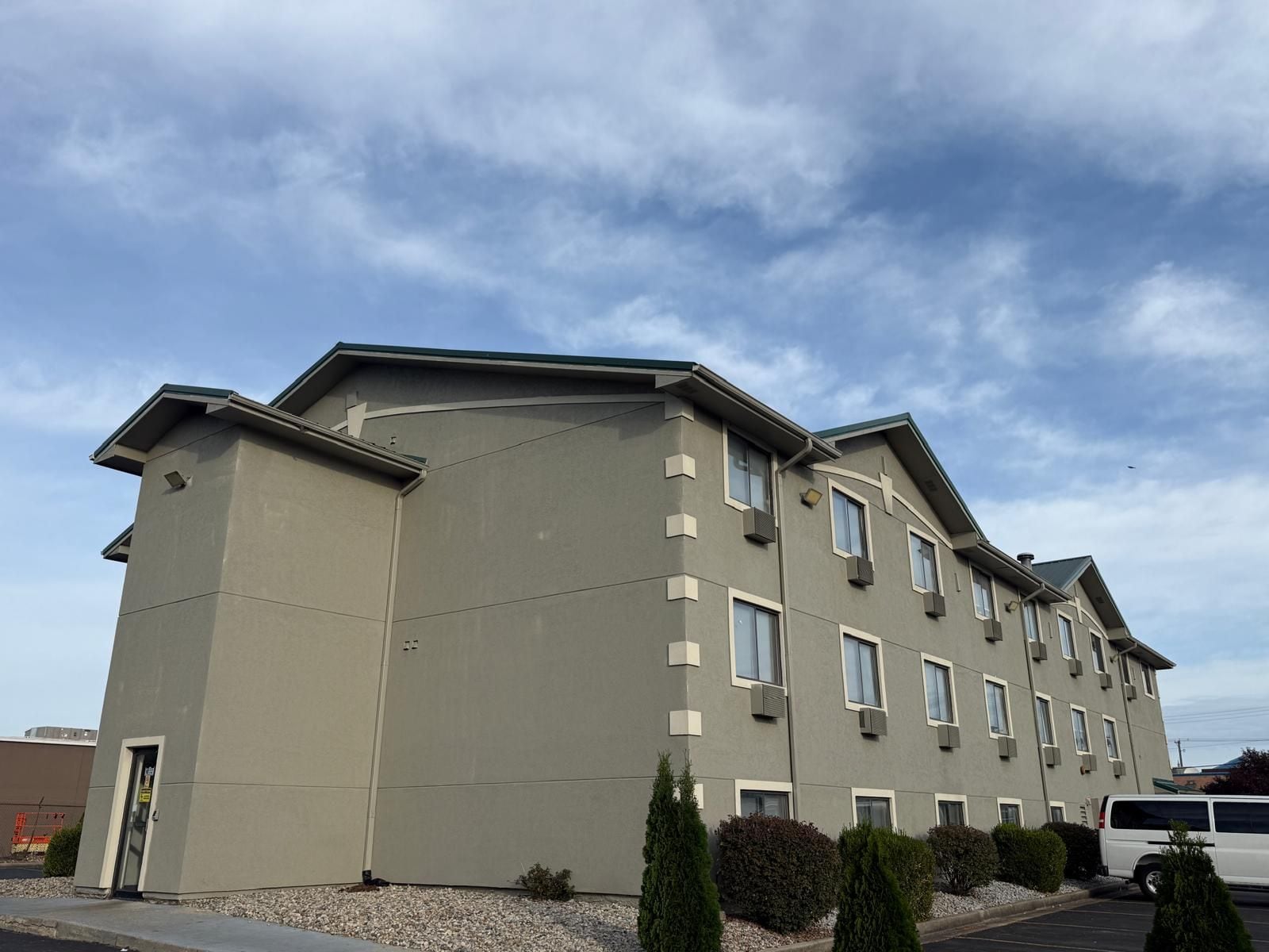 A multi-story hotel with a flat roof, rectangular windows, and a parking area with a white van.