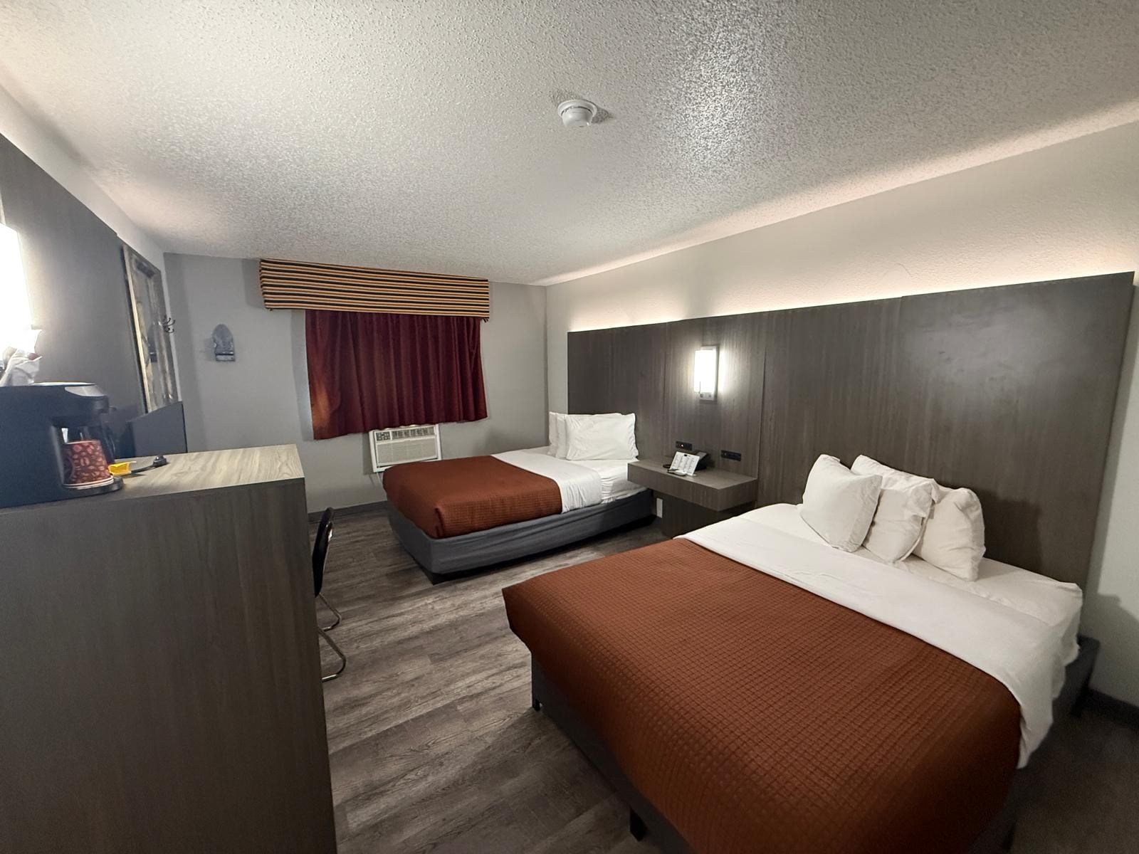 A hotel room with two beds, a desk, a coffee maker, a window with curtains, and a wall-mounted light.