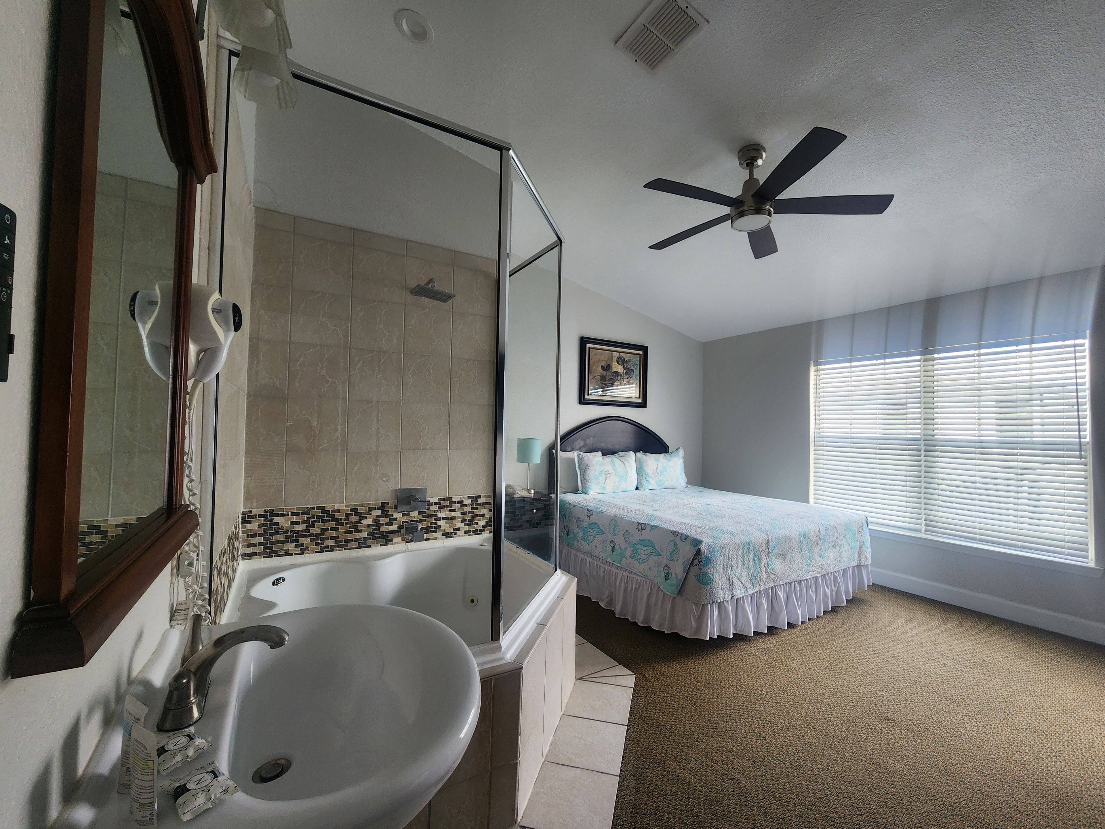 Ein Hotelzimmer mit einer Badewanne, einem großen Spiegel, einem Deckenventilator, einem Bett mit gemustertem Überwurf und einem Fenster mit Jalousien.