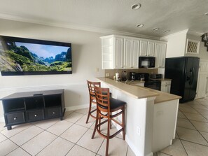 Una cocina moderna con armarios blancos, un refrigerador negro, un microondas integrado y un televisor de pantalla plana grande montado en la pared.