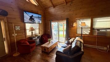 Een gezellig interieur van een blokhut met een houten plafond, een aan de muur bevestigde televisie, een rode fauteuil, een zwarte leren bank en een houten salontafel.
