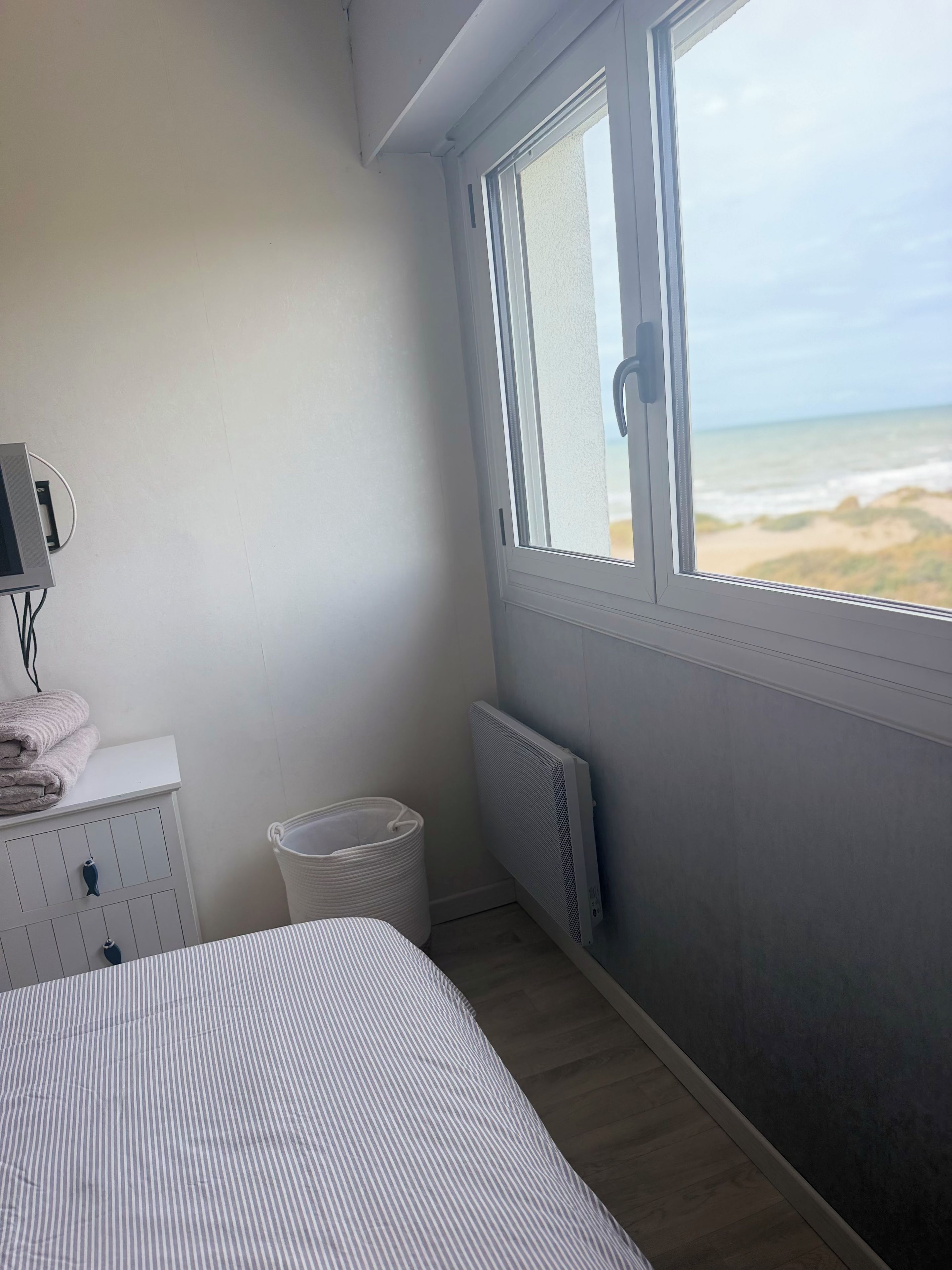 Une chambre avec un lit, un radiateur, une fenêtre donnant sur la plage, une petite commode blanche et une poubelle.