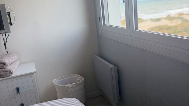 Une chambre avec un lit, un radiateur, une fenêtre donnant sur la plage, une petite commode blanche et une poubelle.