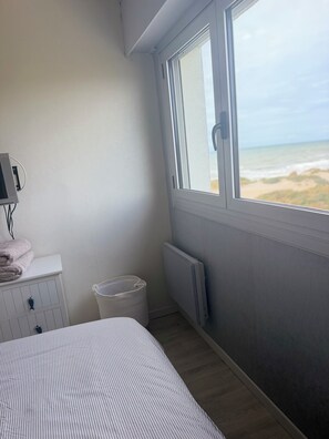 A bedroom with a bed, a radiator, a window with a view of the beach, a small white dresser, and a trash bin. - A gem right by the sea 🌊 the Le Panoramic residence in Sainte-Cécile. (Camiers)