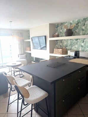A modern kitchen with a central island, bar stools, a flat-screen TV, and a decorative wall with leaf patterns. - A gem right by the sea 🌊 the Le Panoramic residence in Sainte-Cécile. (Camiers)