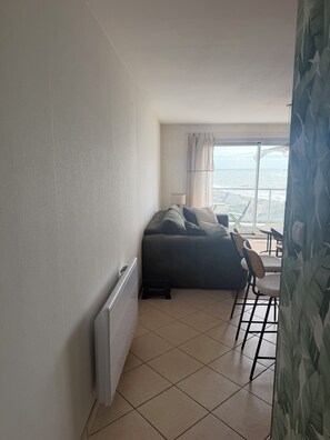 Habitació amb un radiador, un sofà, una cadira i vista al mar a través d'una finestra amb cortines.