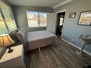 Ein Schlafzimmer mit einem Bett, einem Fenster, einem Ventilator und einer Tür zu einem anderen Raum.