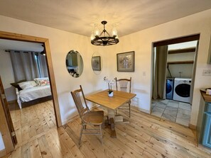 A room with a wooden floor, a dining table with chairs, a chandelier, a bed with curtains, a washing machine, and a mirror on the wall. - Peaceful Farmhouse - Hot Tub - Office (Silt)