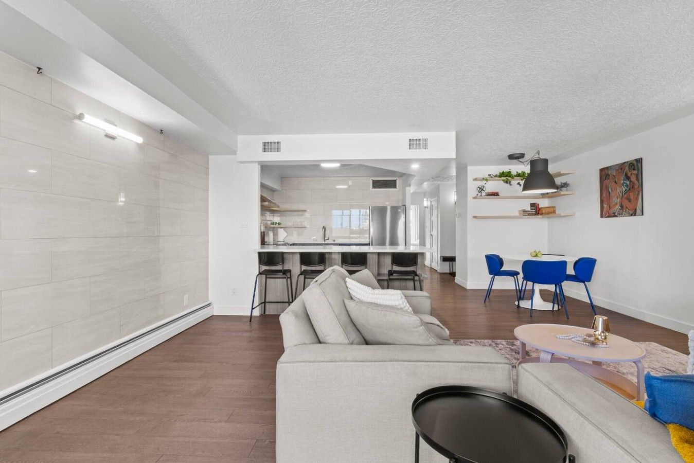A modern living room with a sofa, a dining area with blue chairs, and a kitchen in the background.