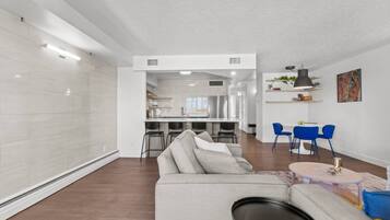 A modern living room with a sofa, a dining area with blue chairs, and a kitchen in the background.
