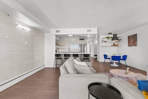 A modern living room with a sofa, a dining area with blue chairs, and a kitchen in the background. - 2BR City Retreat | Stampede, BMO & Free Parking (Calgary)