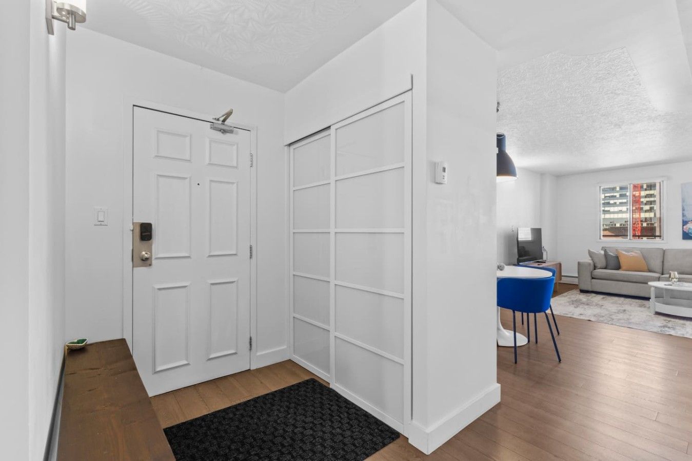 A modern interior with a white door, a sliding closet, a living area with a sofa and a coffee table, and a view of a building outside.