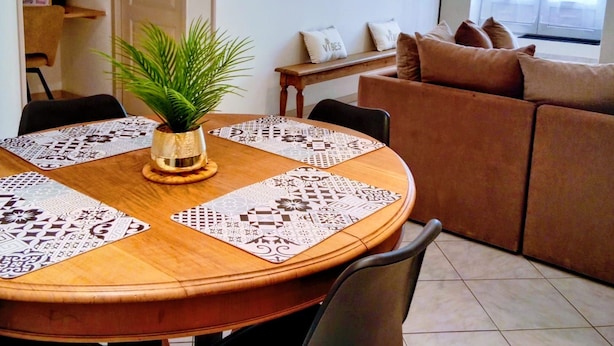 A dining area with a round table, chairs, and a potted plant. There is a sofa and a window with curtains in the background.