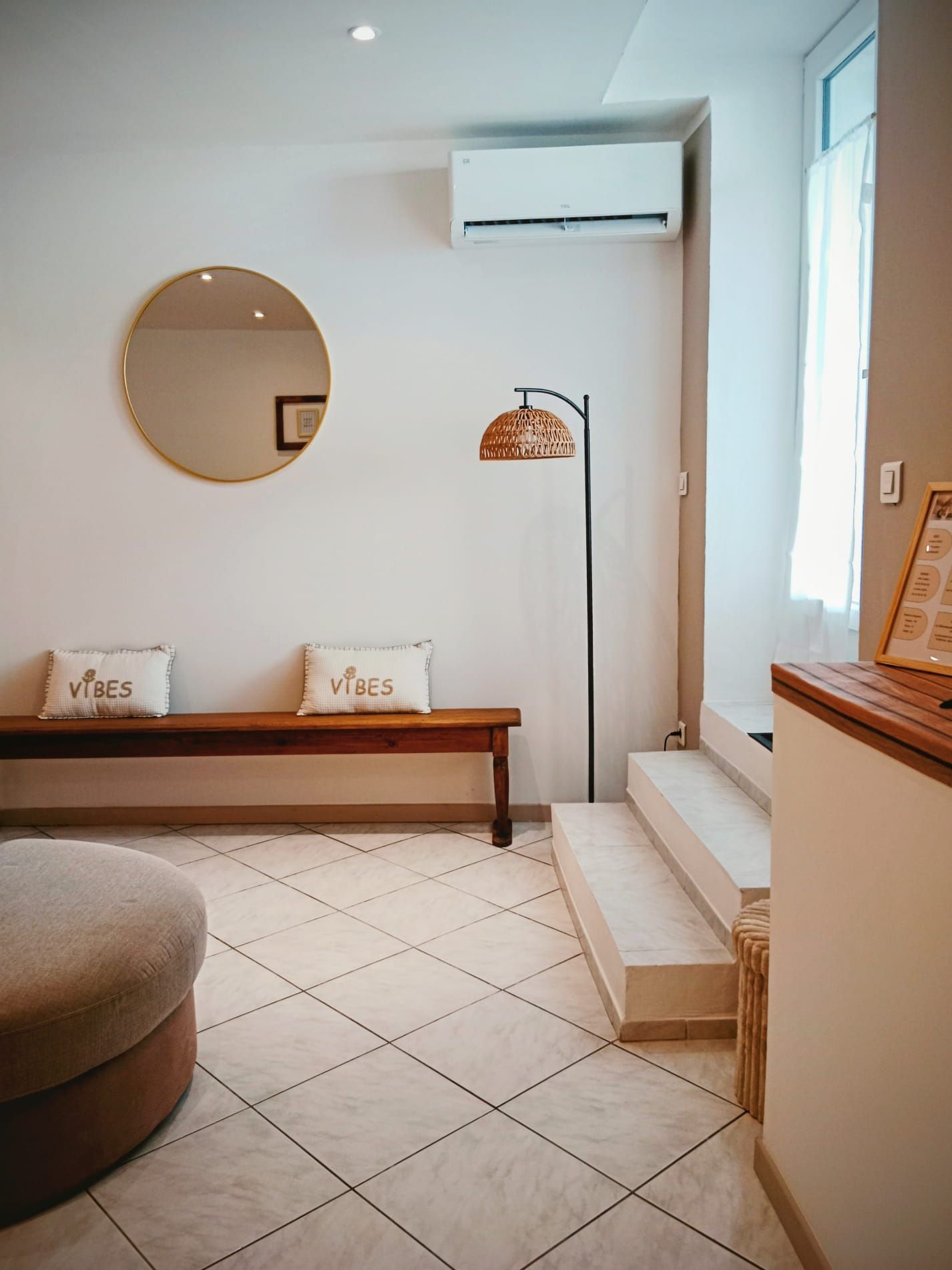 A room with a round mirror, a bench with two pillows, a floor lamp, and a staircase.