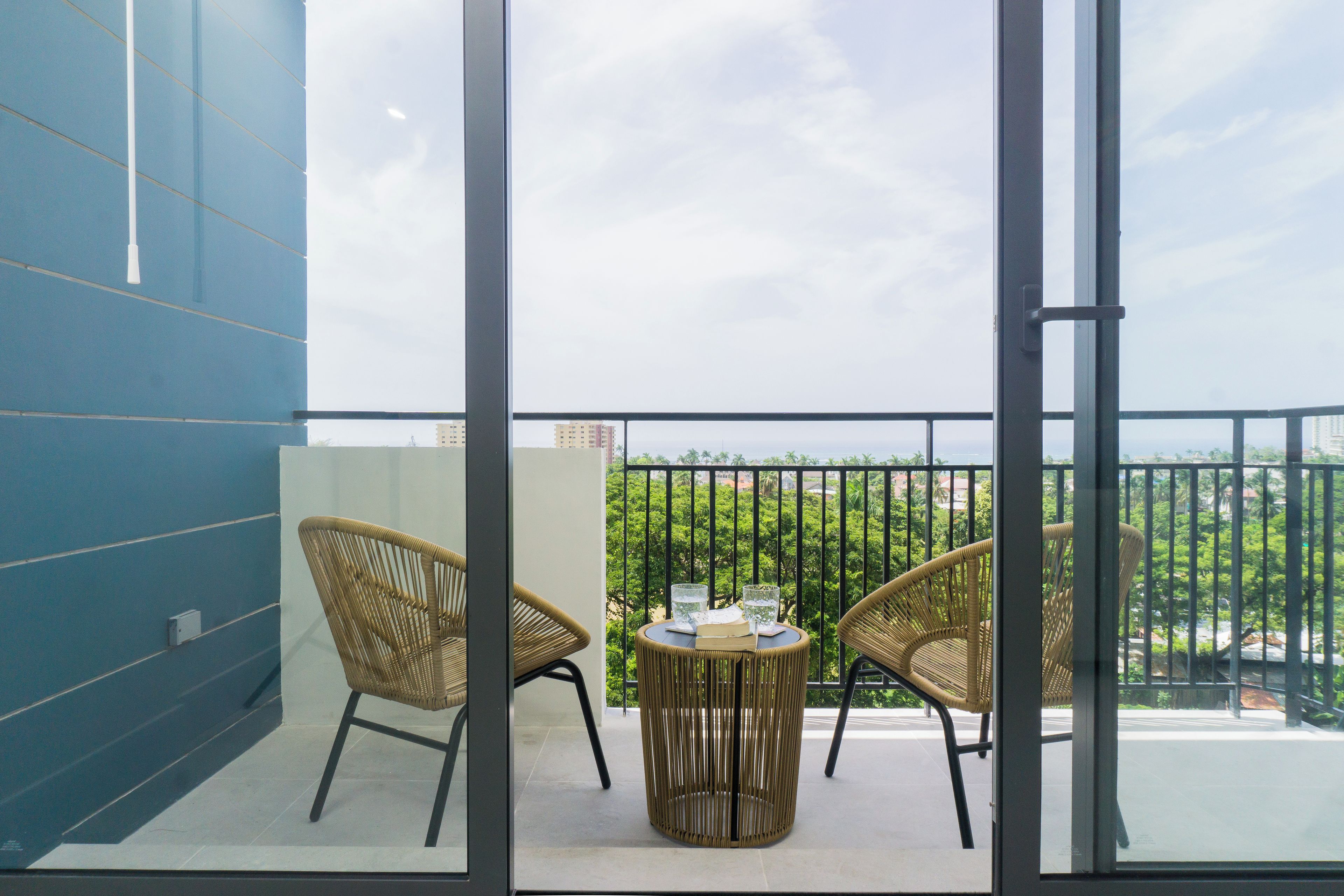 A balcony with two wicker chairs and a small round table, overlooking a green landscape with buildings in the distance.