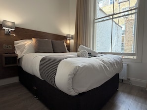 Una cama bien hecha con ropa de cama blanca y un plaid de cuadros, una mesita de noche con una lámpara, y una ventana con vista a un edificio de ladrillo.