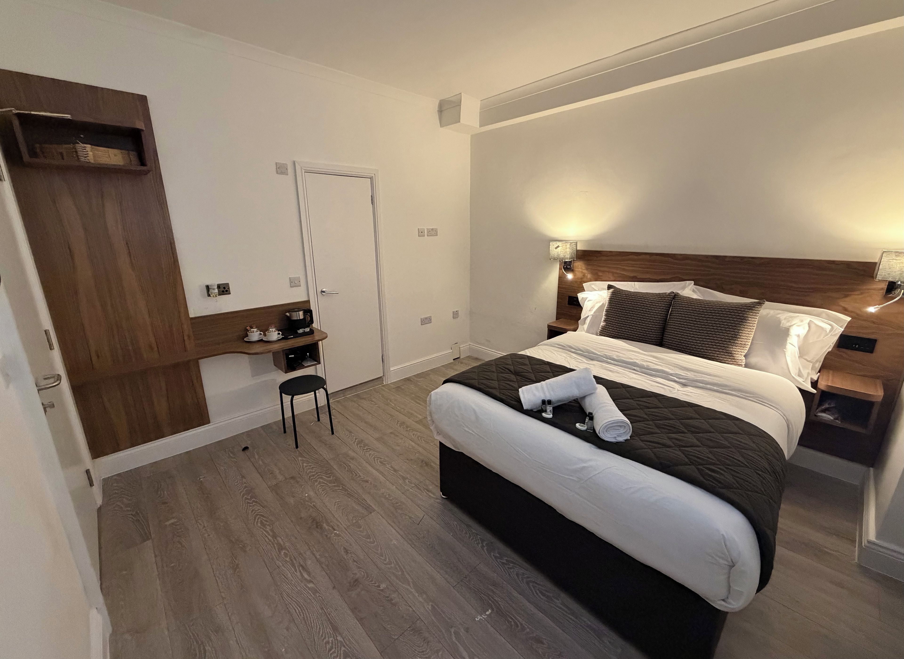 A hotel room with a bed, bedside table, nightstand, and a small desk with a chair.