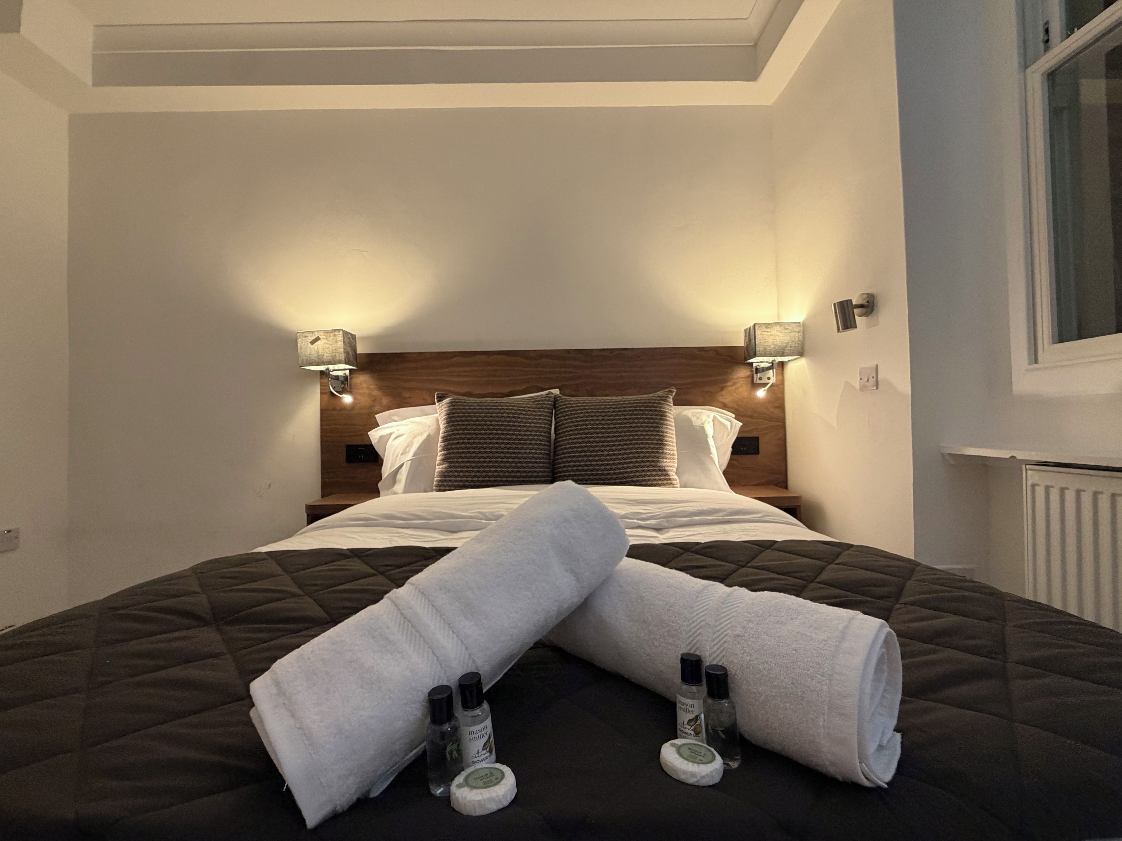 Un lit bien fait, avec une tête de lit matelassée, deux lampes de chevet et une fenêtre sur le côté droit.