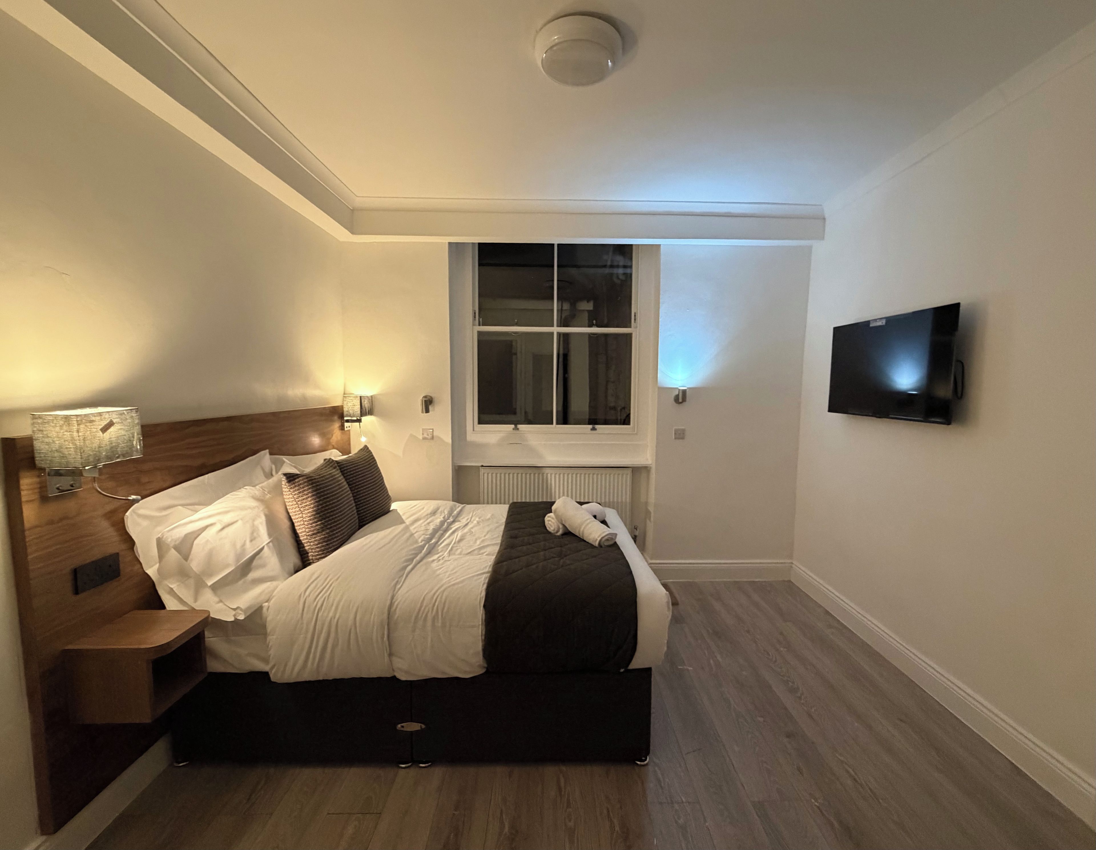 A hotel room with a bed, a nightstand, a lamp, a window, and a television.